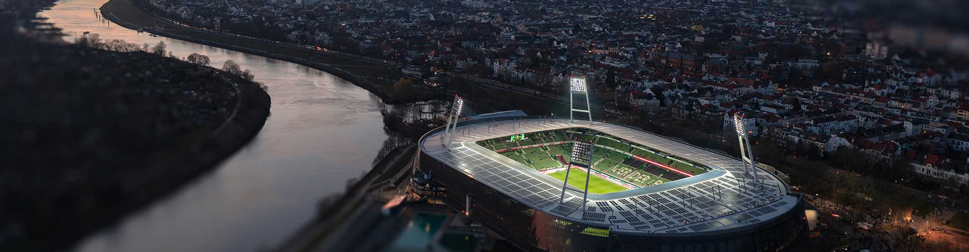 Eine Luftaufnahme vom beleuchteten Weserstadion beim Sonnenuntergang, im Hintergrund ist Bremen zu sehen