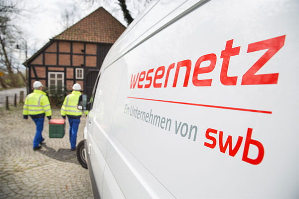 Zwei Mitarbeiter tragen eine Kiste zusammen, im Vordergrund ist auf einem Auto das wesernetz Logo zu lesen