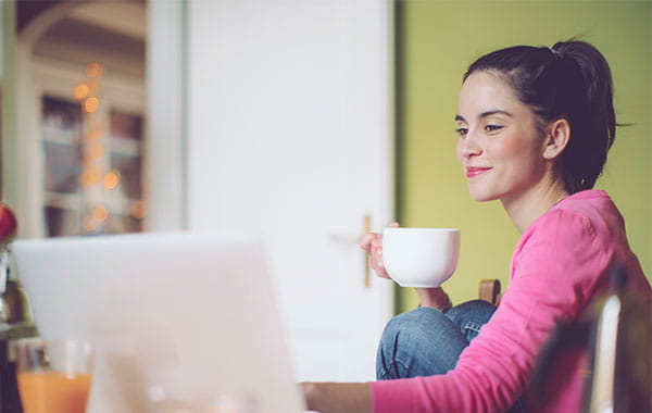 Lächelnde Frau mit weißer Tasse in der Hand schaut entspannt auf Laptop