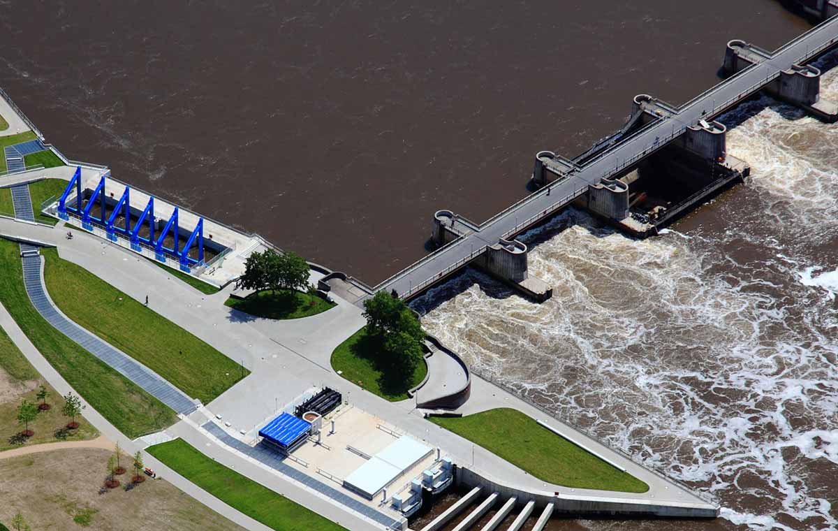 Ein großes Wasserkraftwerk mit einer massiven Staumauer lässt Wasser durch mehrere Schleusen abfließen, umgeben von grünen Bergen und einem breiten Fluss.