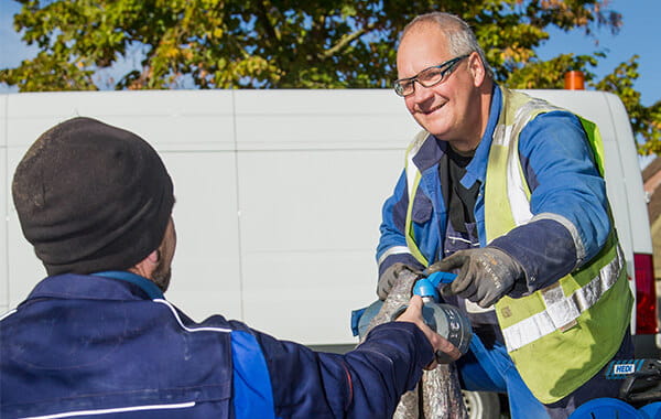 Zwei Mitarbeiter in blauer Arbeitskleidung und Warnweste reichen sich Arbeitsmaterial