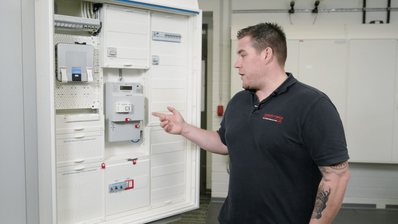 Ein Mitarbeiter zeigt auf einen weißen Zählerschrank. Es ist ein moderner Zählerplatz