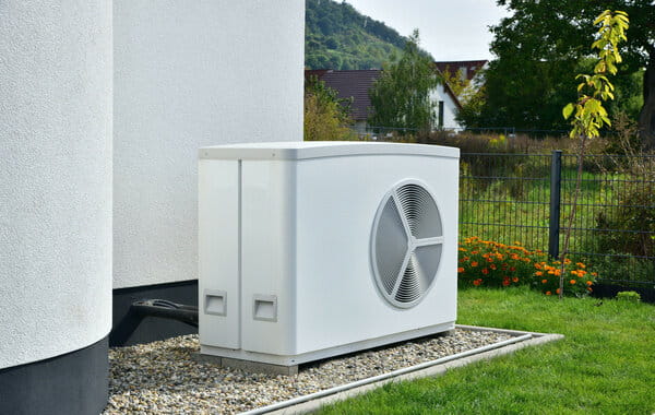 Eine moderne Wärmepumpe steht auf einem Kiesbett neben einem Haus, umgeben von Rasen und Garten.