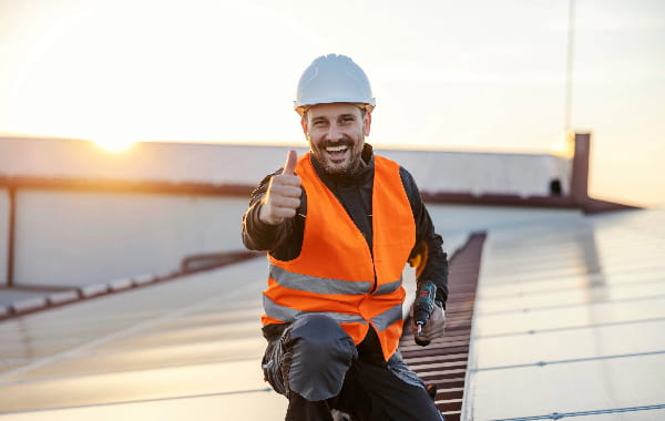 Installateur in orangener Warnweste und Schutzhelm zeigt Daumen hoch auf Dach mit Photovoltaikanlage